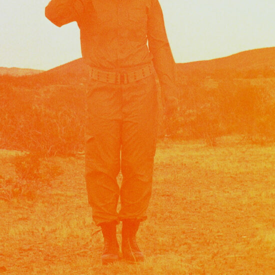 Legs of person in army uniform standing in desert with hills behind. Image is tinted orange.