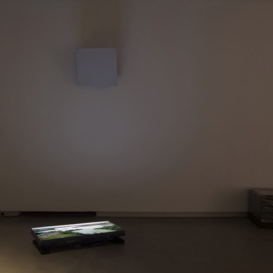 A tv screen lies flat on the concrete floor of Gallery 1, glowing in the dimly lit space and casting a shadow from a square white speaker mounted above it. The bright tv screen displays a green, forested edge of a lake. To its right there is a vinyl wrapped black bench.