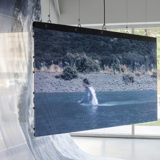 A large flat screen is suspended from the ceiling of Gallery 2, displaying the video documentation of asinnajaq’s performance in a flowing river. In the image it displays, asinnajaq is seen dressed in white trudging through the water, against its current. The riverbank is rocky and occupied by tall grasses and vegetation. Behind the tv screen, an image of a river is printed on nylon, draped from the gallery ceiling and flowing under the video documentation to the floor. In the window behind the piece, a person dressed in yellow can be seen walking past the gallery on the sidewalk.