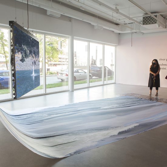 Two people dressed in black are viewing the piece, facing towards the tv screen where asinnajaq walks upstream a rushing river and the river-printed nylon draped behind it. At the feet of the two figures there lies a printed image of wild cranberries. Behind them is a light pink wall that reads “river piece. Submit. Resist”. To the left of the two figures in the gallery window, where cars can be seen parked along the sidewalk.