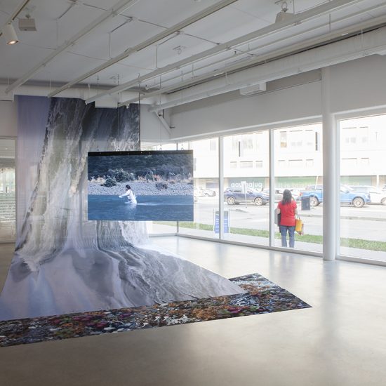 A large flat screen is suspended from the ceiling of Gallery 2, displaying the video documentation of asinnajaq’s performance in a flowing river. In the image it displays, asinnajaq is seen dressed in white trudging through the water, against its current. The riverbank is rocky and occupied by tall grasses and vegetation. Behind the tv screen, an image of a river is printed on nylon, draped from the gallery ceiling and flowing under the video documentation to the floor. In the window to the right of the piece, a person dressed in red and blue can be seen standing, facing away from the camera.