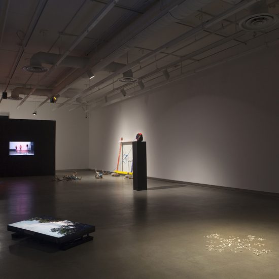 A view from the northeast corner of a dimly lit Gallery 1. Kite’s work, Iron Road comprises the foreground: to the right lies a tv screen flat on the floor, displaying trees against a blue sky, and to the right there are many small rocks, spotlit and arranged in a geometric pattern. Dangers’ work occupies the background: a tv screen mounted on a black wall, displaying two figures cast in red light. To the right of the screen there is remaining ephemera from Danger’s performance- skulls and antlers connected by blue rope, four pieces of wood joined to form a white, red, yellow, and blue frame that leans against the gallery wall, and a red and black beaded mask on a tall pedestal.