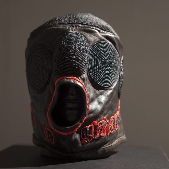 A leather mask adorned with red and black beads sits on a black pedestal, spotlit from the right. The mask covers the entirety of the mannequin head it rests on, save for an opening around the mouth that is outlined by a circle of red beads. There are black beaded circles orbiting the top of the mask, and lower, around the neck, there is black and red beaded lettering.