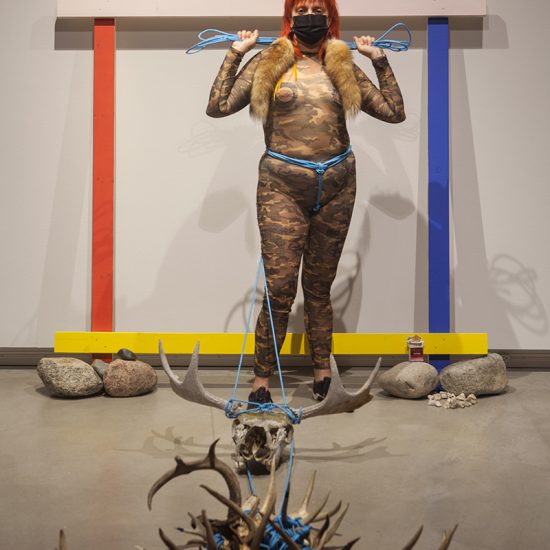 Danger stands proudly, holding a bundle of bright blue rope tightly above their shoulders in front of the red, pink, blue, and yellow frame. The frame is flanked by two piles of rocks- one which supports a pack of cigarettes. In the foreground there are skulls and antlers, tethered to Danger’s waist by the blue rope.