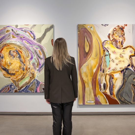 A person with blonde hair wearing a green blazer stands in front of two paintings by Manuel Mathieu, hung on a light blue wall.