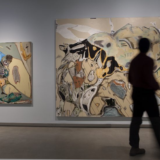 A person walks by two paintings hung on a light blue wall. The painting on the left is smaller than the one on the right, which takes up most of the wall.