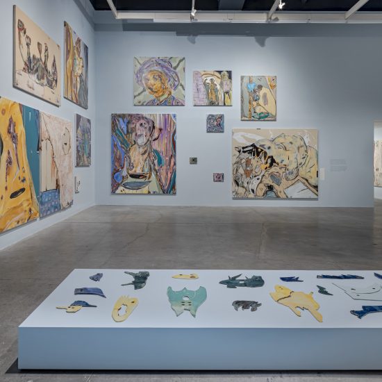 An array of paintings by Manuel Mathieu are displayed in the corner of a gallery at The Power Plant Contemporary Art Gallery in Toronto. The walls are painted a soft bay blue. In the foreground of the image, a matching blue plinth, low to the ground, displays ceramic works by Mathieu in dark blue, turquoise and yellow.