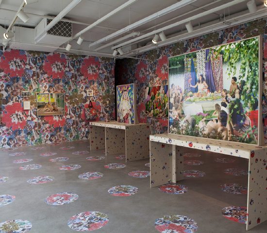 Photographs by Leonard Suryajaya hung on gallery walls that are wallpapered with a collage. Some stand on tables decorated with stickers of the same design. The floor has a circular checkerboard pattern of the collage.