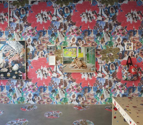 Two photographs by Leonard Suryajaya hung on gallery wall. The wall is covered in a wallpapered collage and there is a tote hanging on the wall with the same pattern. The corner of a table is visible with a lucky cat on top of it.