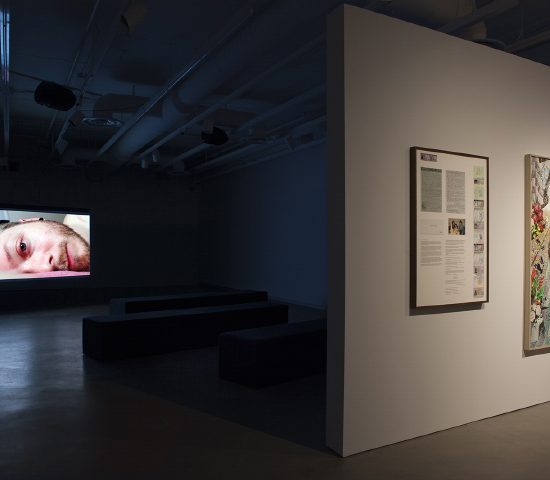 Room with video installation showing a man’s face on screen and a partitioned wall with two photographs hanging on it