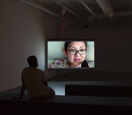 Person sitting on bench and watching the video installation and sit on a bench. A woman’s face is on screen and she’s speaking into a microphone.