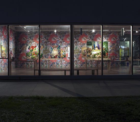 View of gallery at night from the street, captured through wall of gallery windows. The sidewalk and grass are visible.
