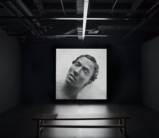 Dark gallery space with an image of a face in black and white projected, there is a bench directly in front for viewing