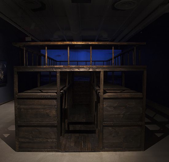 Dark room with projection of water on the back wall, a large slave ship is in the middle of the room