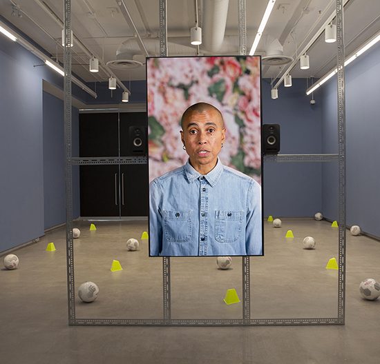 Room with soccer balls and cones on the ground, in the foreground a metal frame with large tv screen with a woman's face on it