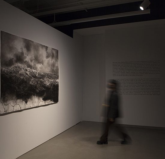 A blurred figure stands between a large charcoal drawing and large piece of text printed on the wall