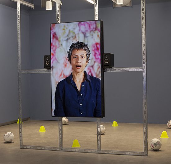 Room with soccer balls and cones scattered on the floor. A large metal frame stands holding a tv screen playing a video of a woman singing
