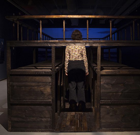 Woman stands in the middle of wooden ship, projection of water plays on the back wall