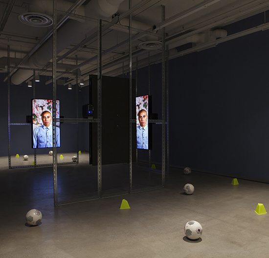 4 large metal frames stand in the middle of the room with tv screens hanging on each of them. Soccer balls and cones are scattered around the outside of the structure