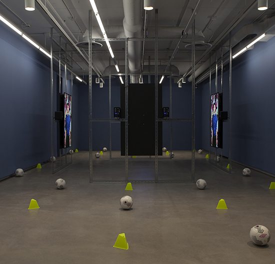 Room with soccer balls and cones are scattered around a metal square structure in the middle of the room. Each frame has a large tv screen hanging on it