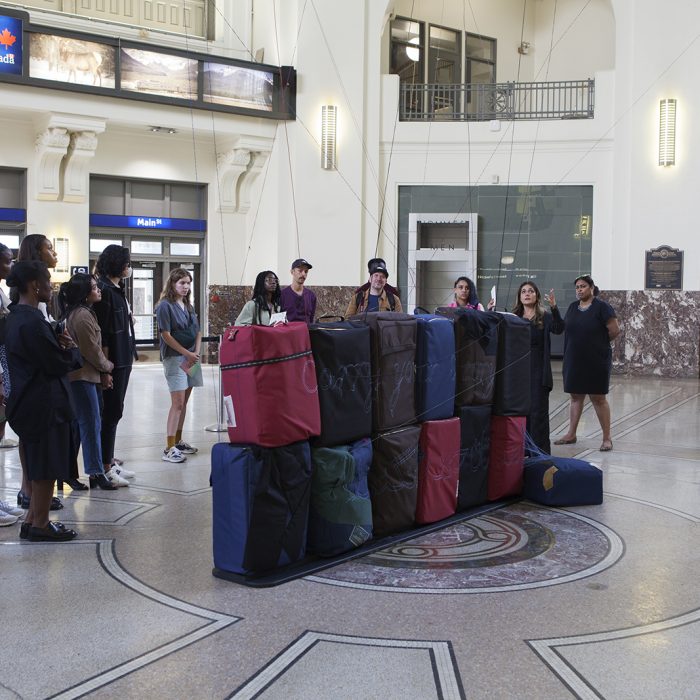 A group of people stand to the left side of the image watching and listening to a speaker. They are gathered around several soft sculptures that resemble luggage.