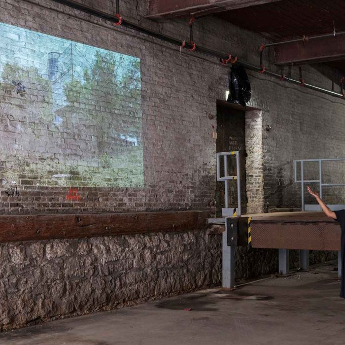 A video projected onto a white brick wall. The artist Marisa Gallemit stands in front of it and gestures to it.