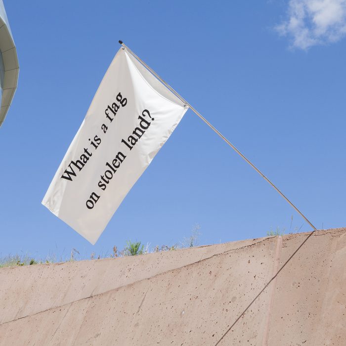 A white flag mounted to a beige stone wall. The flag has black text on it that reads "What is a flag on stolen land?"