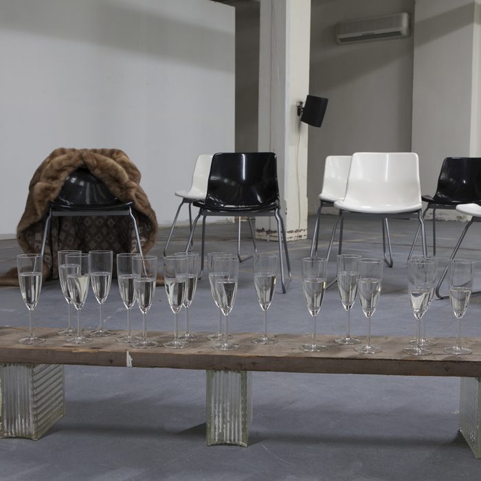 Several champagne flutes filled with water sit on a wooden bench on a grey floor. In the background there are many black and white chairs, one of which has a brown fur coat draped on it.