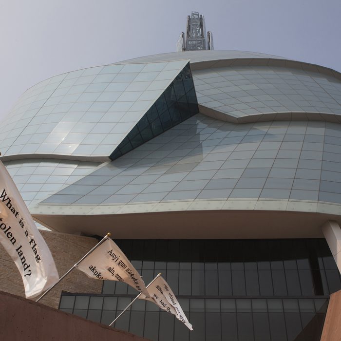 Three white flags with black text flying along the wall of the Canadian Museum for Human Rights.