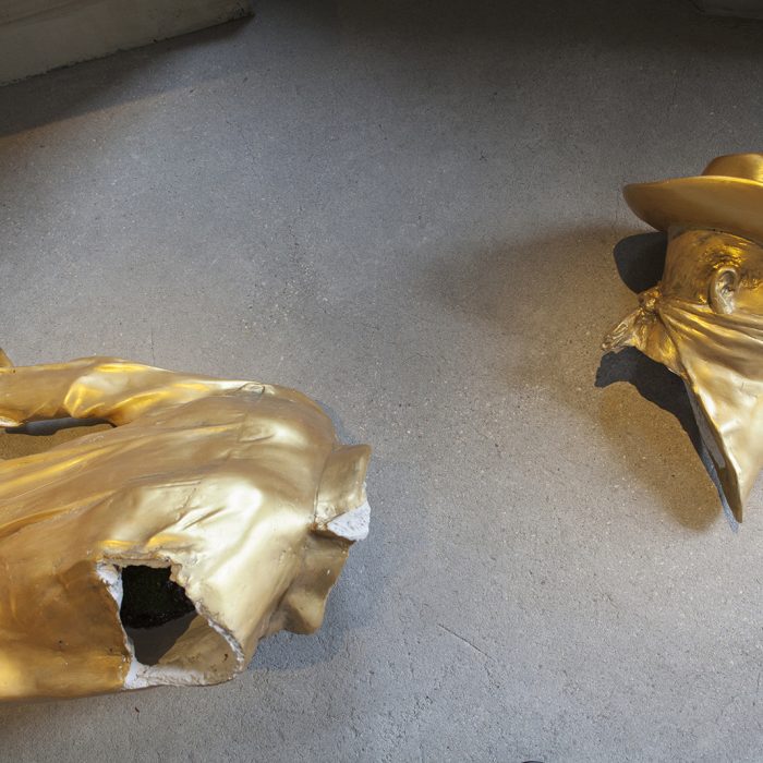 The head and torso of a golden statue lie broken on the ground. In the hollows of its fractured form, one can see mushrooms and grasses growing.
