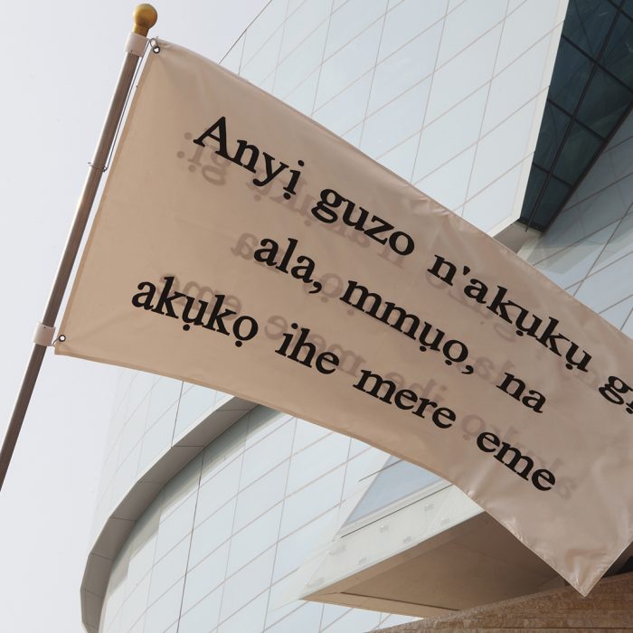 A close-up of a white flag with black text flying in front of the Canadian Museum of Human Rights. The flag reads a phrase in Igbo Anyị na-eguzo n'akụkụ gị: ala, mmụọ na akụkọ ihe mere eme