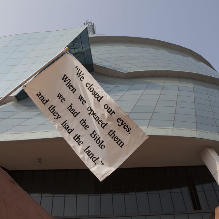 A close-up of a white flag with black text flying in front of the Canadian Museum of Human Rights. The flag reads We closed our eyes. WHen we opened them we had the Bible and they had the land. In English
