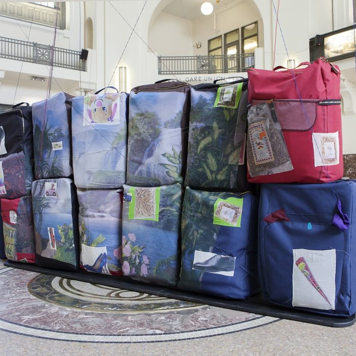 Several soft sculptures that resemble luggage are stacked together. Landscapes, cartoon characters, and other images are printed and embroidered onto them.