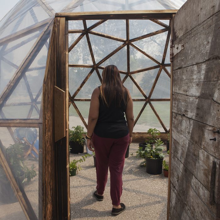 A dome with a wooden frame covered with clear sheeting. The door to the dome is open and there are many plants in pots within it. We watch a person walking into the structure.