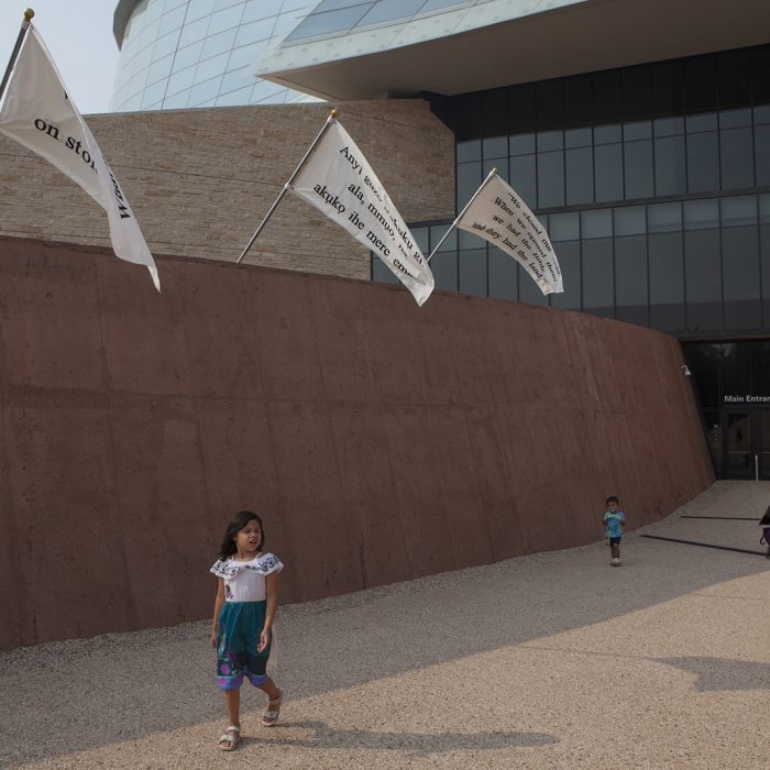 Three white flags with black text flying along the wall of the Canadian Museum for Human Rights. A child walks towards the camera in front of the wall, with their family trailing further in the background.