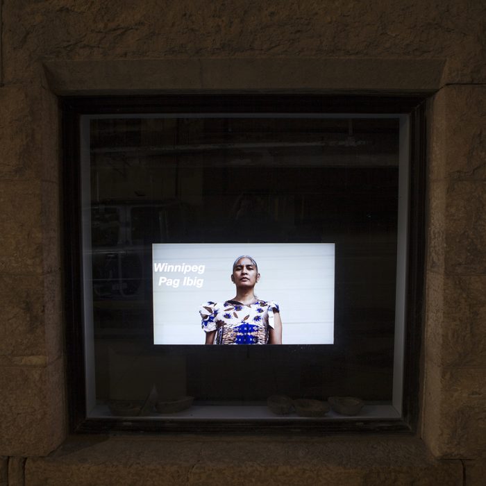A video displayed in a window. Halved coconut shells sit on the inside window shelf in front of the screen. The text on the video screen says "Winnipeg Pag Ibig" and pictures a woman from the ribs up wearing a purple dress and looking directly at the camera.