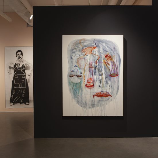 a blue, pink,purple abstract painting hangs on a black wall in the foreground. In the background a light pink wall can be seen with a large photo of a woman hanging on it.