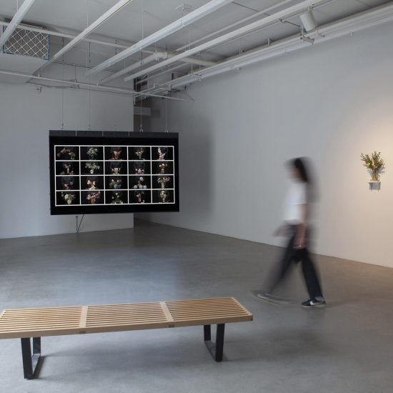 Interior gallery 2, a woman walks through the middle of the gallery. In the foreground a bench, to the back left a large screen hangs, on the right a glass boot holds a small bouquet of flowers on a small shelf