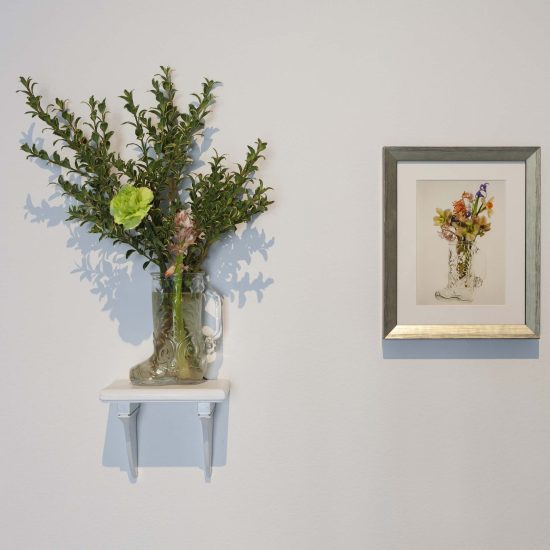 A glass boot mug holds a small bouquet of flowers on sits on a small shelf. To the right, a small framed photo of the bouquet and boot hangs in a silver frame.