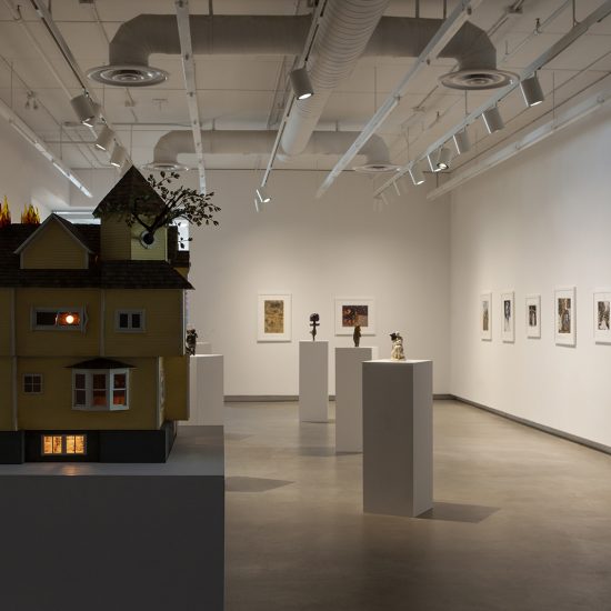 Point o f view from back wall of gallery one facing the entrance. House on Fire is large in the foreground. Subsequent five podiums containing smaller statues in the back. Photos hung on the right and back wall.