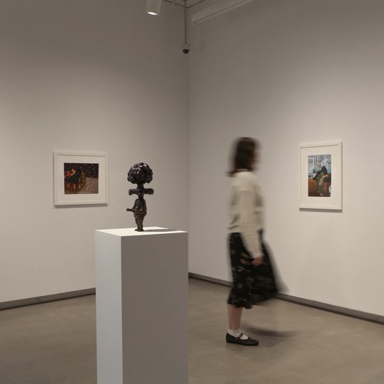Rectangular white podium with small brass statue in the foreground. Woman engages with a picture on the wall to the right. Another picture hangs on the back wall to the left.