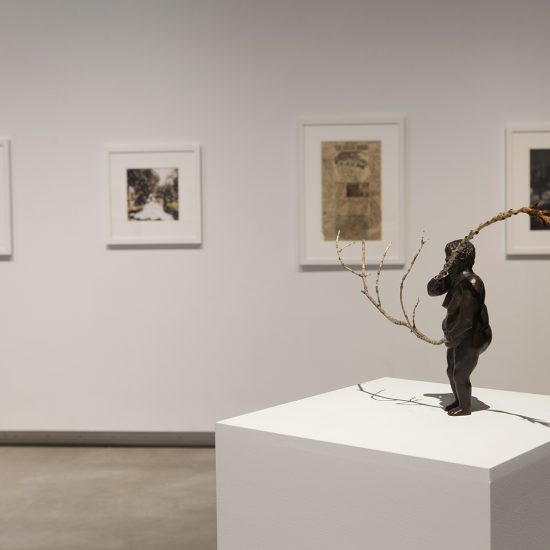 Rectangular white podium sits in the foreground. On top is a bronze figure statue with twigs extending from the arms. Behind pictures hang on wall in line.