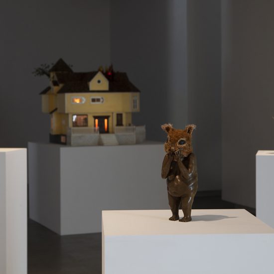 Rectangular white podium in foreground showcasing mixed media figure statue. The body is bronze with a squirrel head, the arms meet up as the hands cover the mouth. In the back round sits the large yellow dollhouse lit up from within.