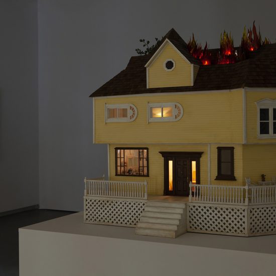 Close up of House on Fire. The yellow, white trimmed wooden dollhouse sits in a shadow of the gallery. This allows for the lights inside and the fire on the roof to be seen.